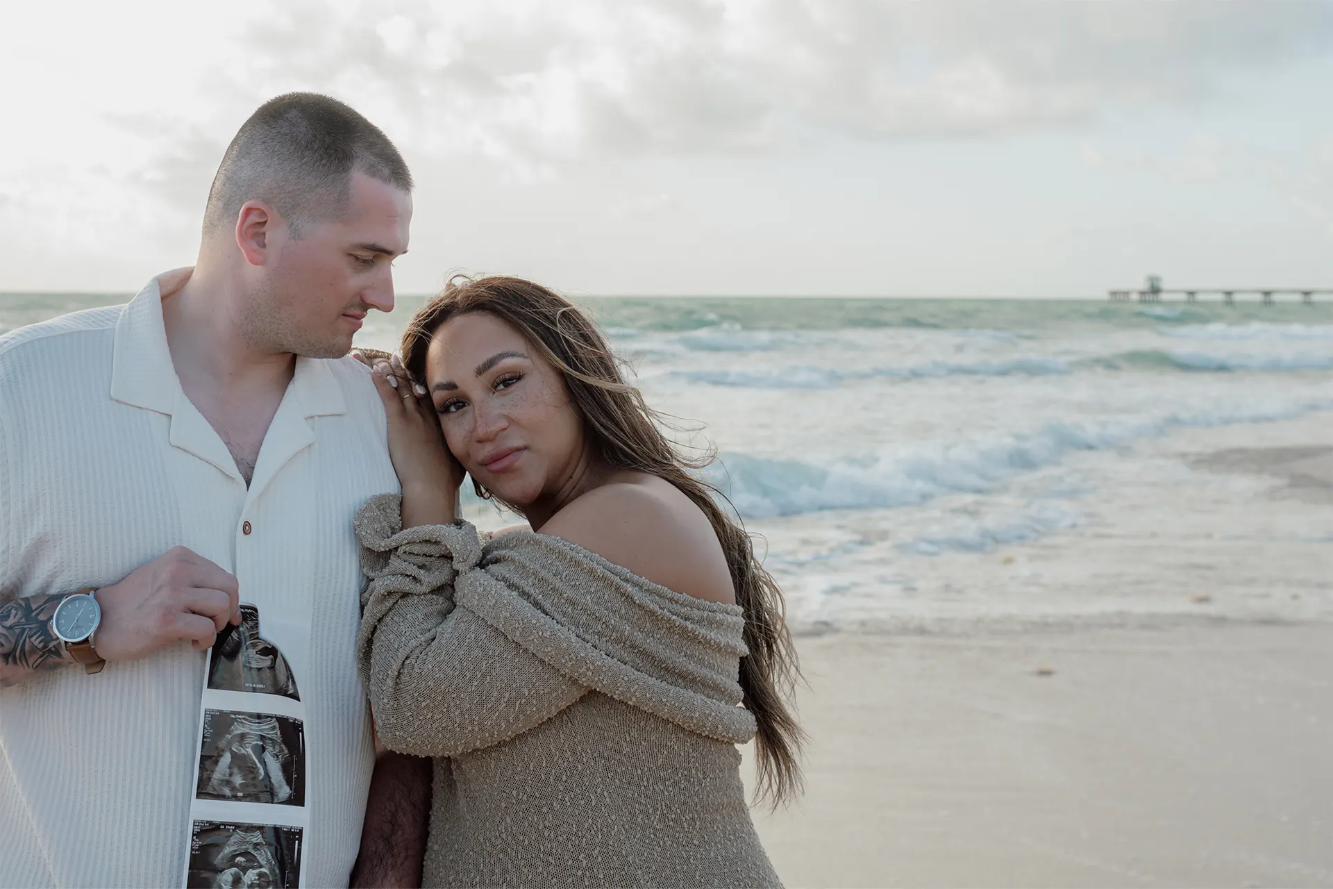 Expecting couple standing barefoot on the sand holding their baby sonogram