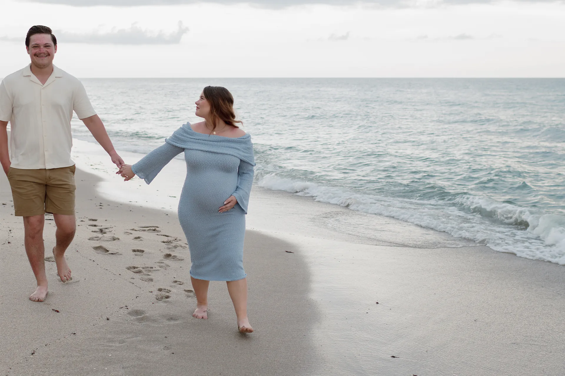 Maternity photoshoot at the beach featuring expecting parents walking on the sand