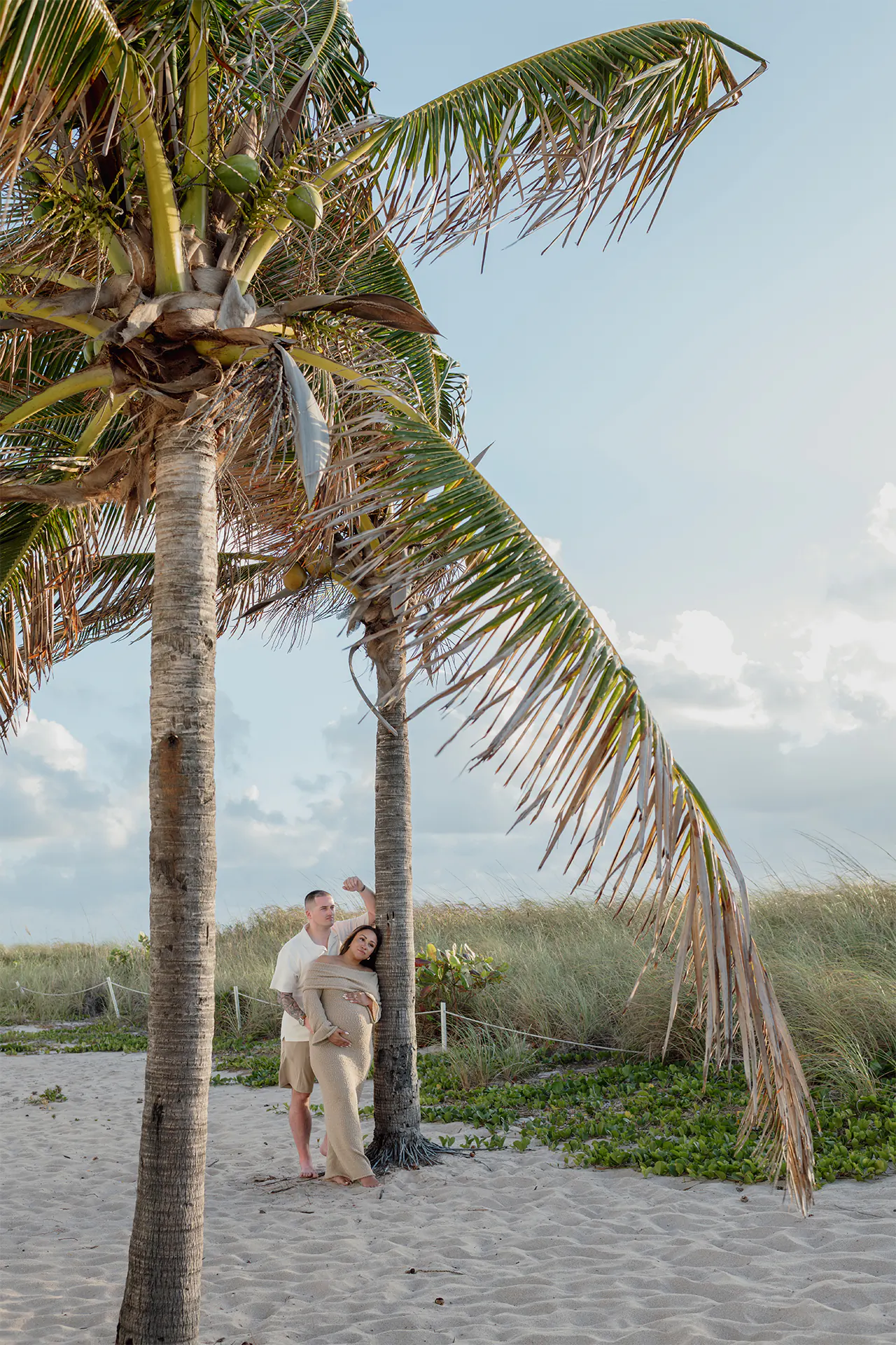 Expecting couple Alex and Todd leaning on a palm tree during their beach session