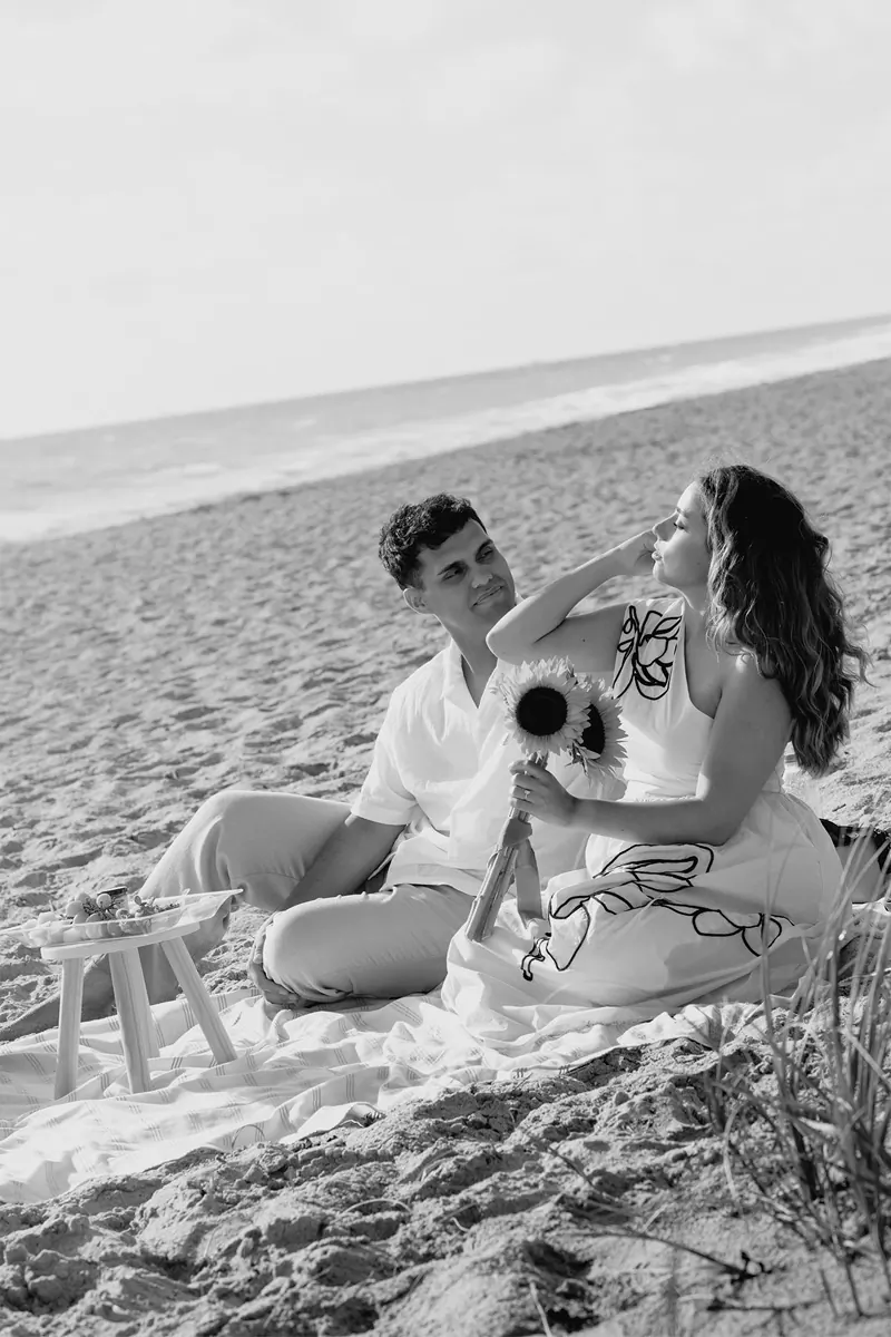 Black and white image of a couple sitting down on the beach after their marriage proposal